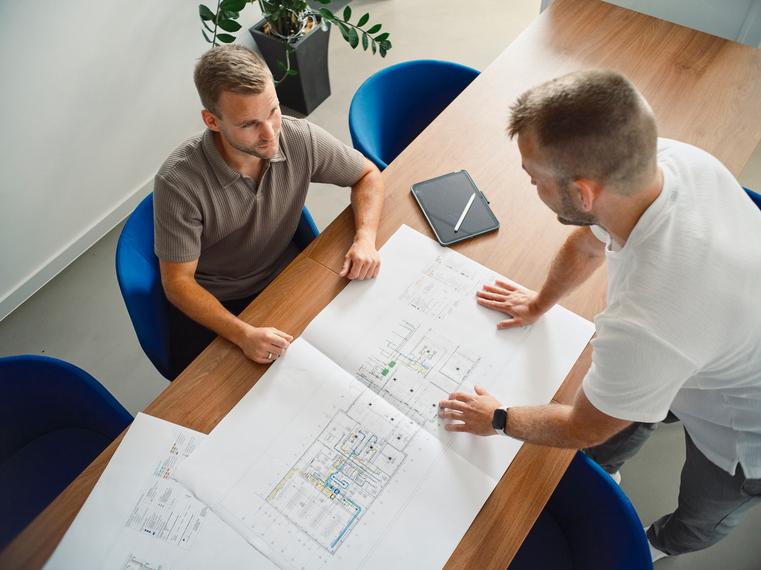 Zwei Männer stehen über Bauplänen an einem Tisch und besprechen Details.