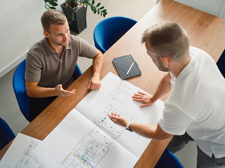 Zwei Männer stehen über Bauplänen an einem Tisch und besprechen Details.