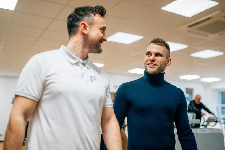 Zwei Männer gehen lächelnd durch ein Fitnessstudio, im Hintergrund Sportgeräte.