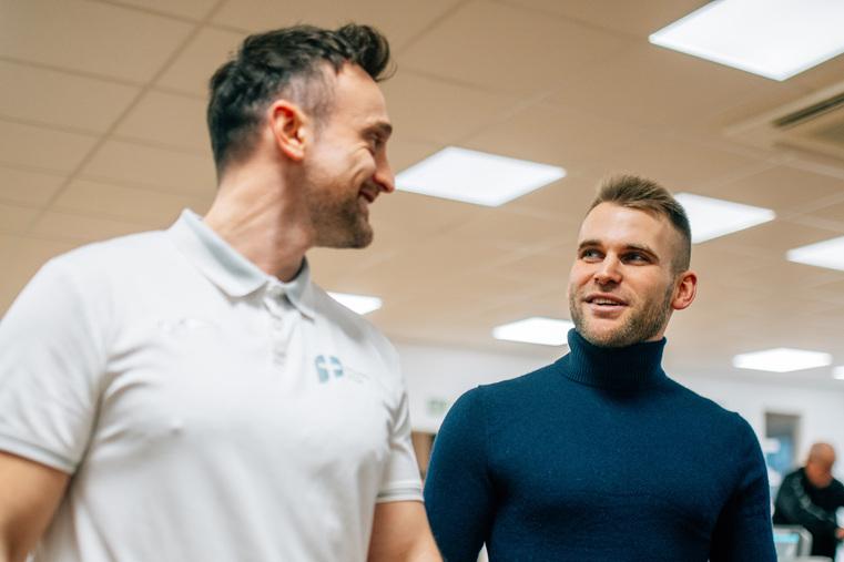 Zwei Männer gehen lächelnd durch ein Fitnessstudio, im Hintergrund Sportgeräte.