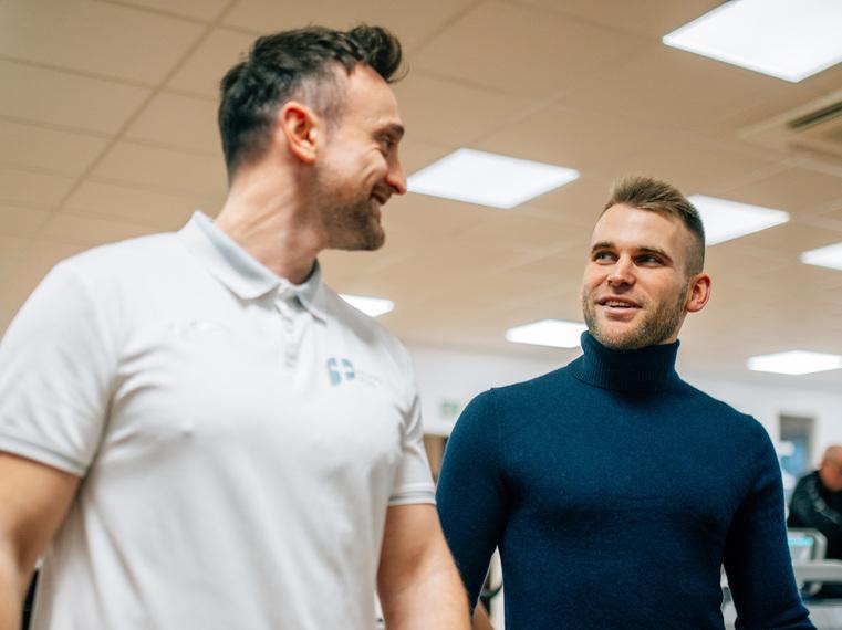 Zwei Männer gehen lächelnd durch ein Fitnessstudio, im Hintergrund Sportgeräte.