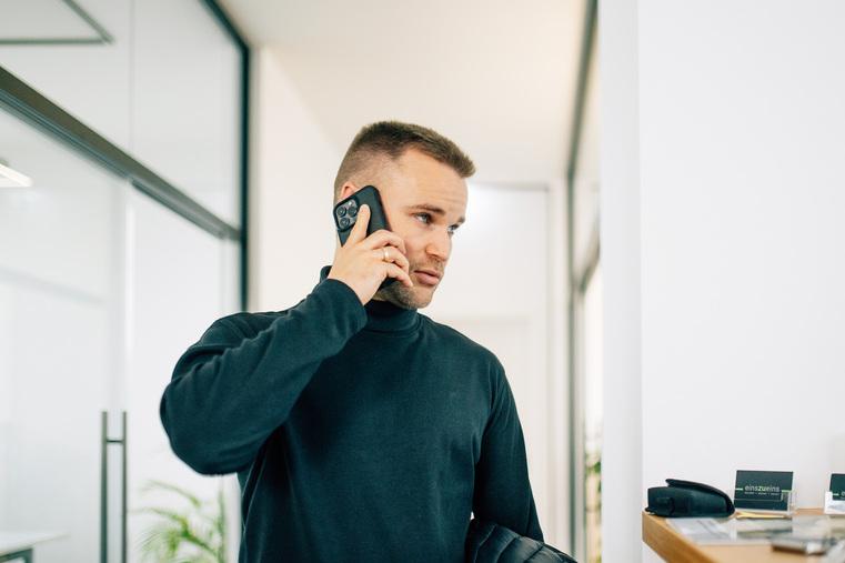 Mann in dunklem Rollkragenpullover telefoniert mit Smartphone in einem hellen Büroflur.