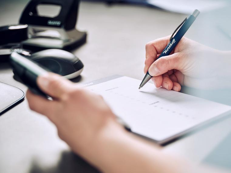 Eine Hand schreibt mit einem "Northoff" Kugelschreiber auf einem Notizblock, daneben ein Smartphone.
