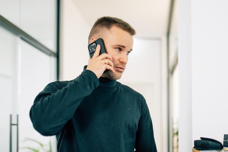 Mann in dunklem Rollkragenpullover telefoniert mit Smartphone in einem hellen Büroflur.