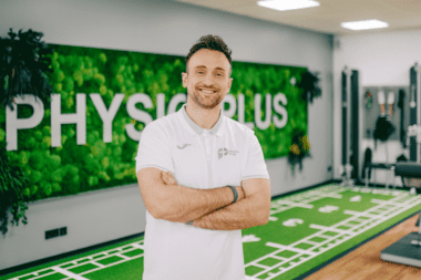 Trainer lächelt in Physiotherapie-Praxis mit Kunstrasen. Stellt Klimasysteme und Lüftungstechnik für Sportstätten dar.