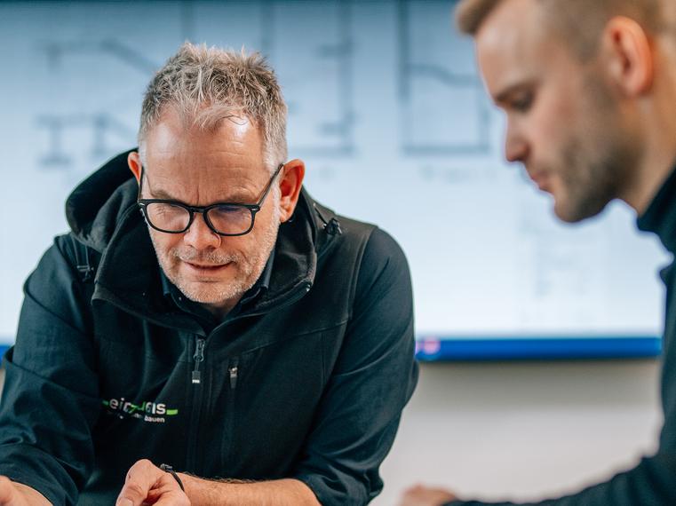 Zwei Männer in Arbeitskleidung blicken auf Planungsunterlagen, ein Bildschirm mit Bauplänen im Hintergrund.